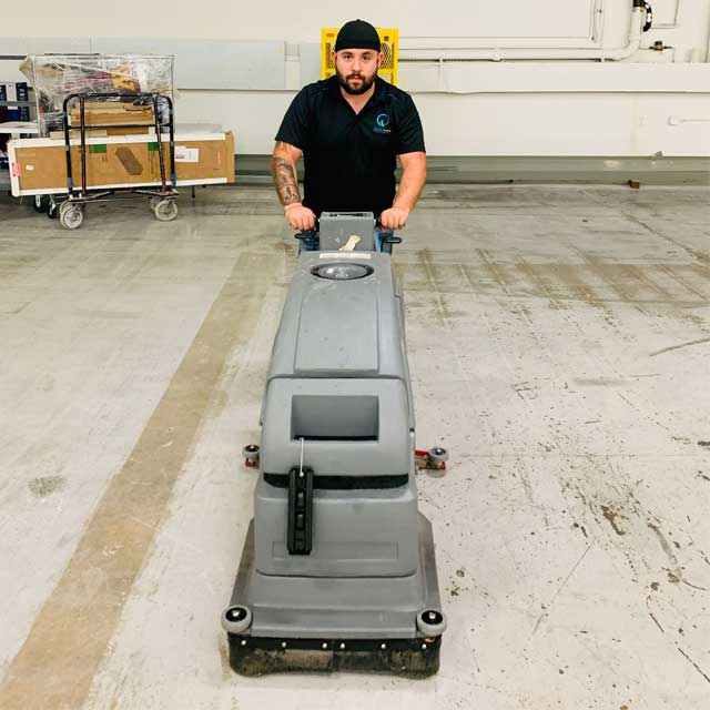 A person wearing a black shirt and cap pushes a large floor cleaning machine across a spacious, empty warehouse floor, representing efficient commercial cleaning services. In the background, a cart with boxes complements the well-lit industrial scene, showcasing the thoroughness of Latin Group Commercial Cleaning.