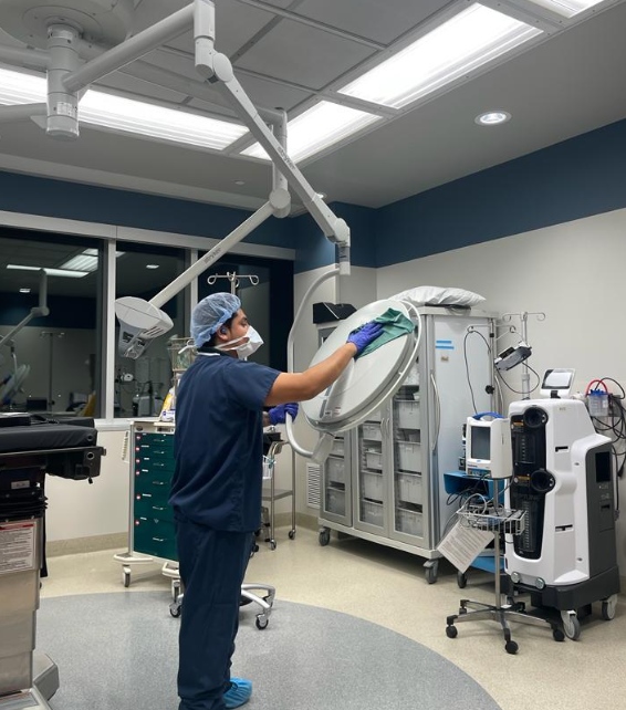 A medical professional in scrubs and a hairnet adjusts an overhead surgical light in a spotless hospital operating room, maintained by Latin Group Commercial Cleaning. The room is equipped with medical supplies, cabinets, and various medical devices.