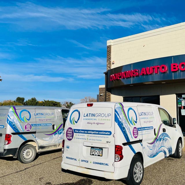 Two white vans, adorned with "Latin Group Commercial Cleaning" branding, are stationed outside a building marked "HOPKINS AUTO BODY." The vibrant graphics and contact details highlight their premier cleaning services. Above, a clear blue sky sets the perfect backdrop.