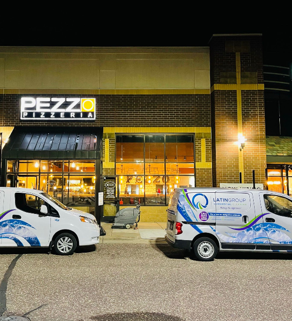 Two white vans parked in front of Pezzo Pizzeria, a brick building with a lit sign, showcase their vibrant decals promoting Latin Group Commercial Cleaning. Parked on a street at night, the restaurant's large windows glow warmly, reflecting the commitment to pristine spaces by top-notch cleaning services.