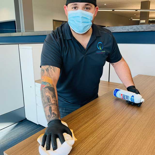 A person from Latin Group Commercial Cleaning, wearing a mask and gloves, efficiently cleans a wooden table with a cloth and cleaning spray. Sporting a tattoo on their arm and dressed in a dark polo shirt and cap, they seem to be tidying up an office or commercial space.