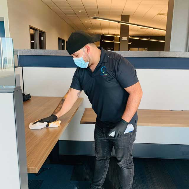 A person wearing a mask, cap, and gloves is diligently cleaning a desk with a cloth and spray bottle, embodying the professionalism of Latin Group Commercial Cleaning. The room features cubicles and ceiling lighting, highlighting the efficiency of top-tier commercial cleaning services.