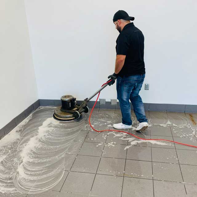 A person from Latin Group Commercial Cleaning is operating a floor buffer to clean tile flooring in a room with white walls. They wear a black shirt, jeans, gloves, and a cap. Soap suds trail the machine's path, showcasing their expert cleaning services.