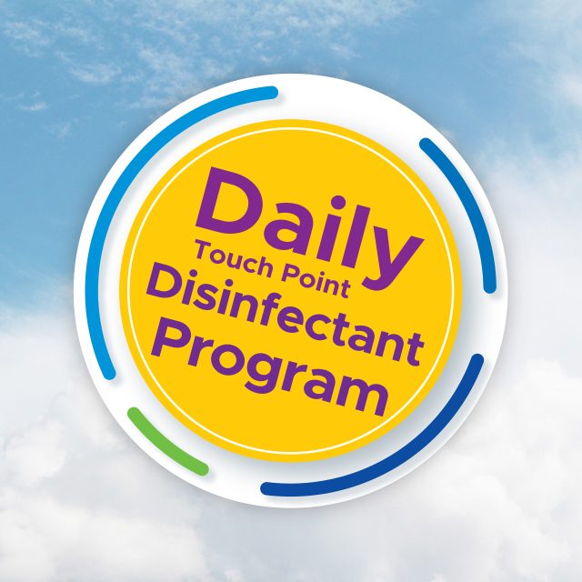 Yellow circular sign with "Daily Touch Point Disinfectant Program" written in purple. The sign, featuring blue and green accents, promotes our Latin Group Commercial Cleaning services against a backdrop of a cloudy blue sky.