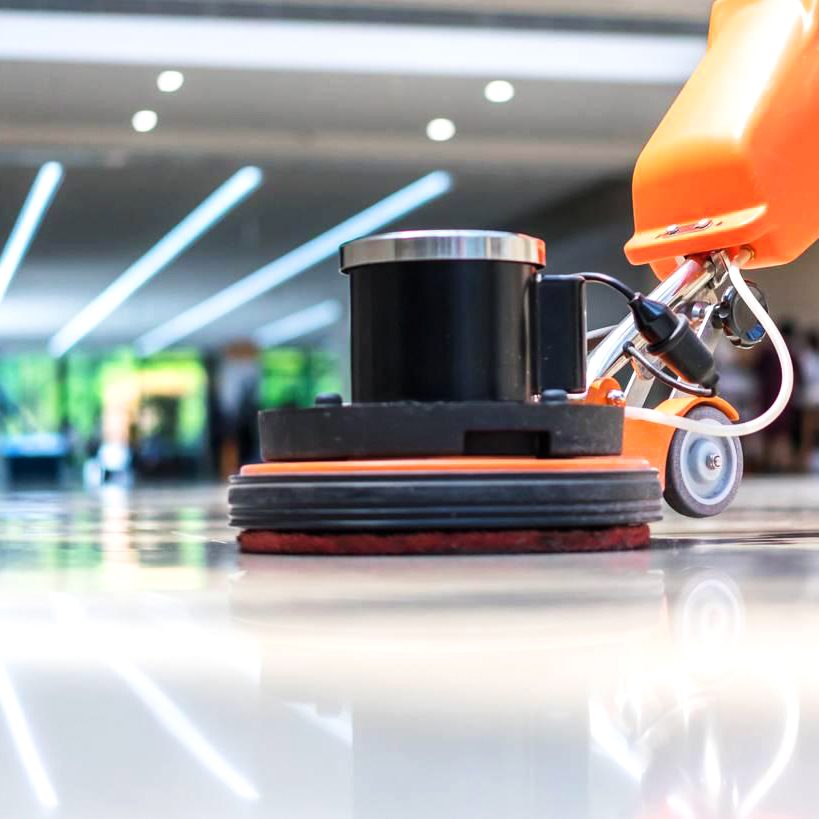 A close-up of an orange floor polishing machine in action on a shiny, reflective surface, showcasing the excellence of commercial cleaning services. The background is slightly blurred, revealing a modern room with bright lighting, indicative of Latin Group Commercial Cleaning's professional touch.