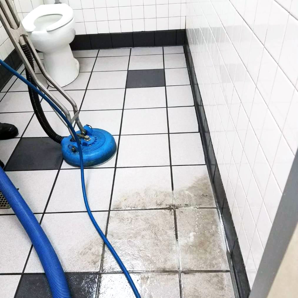 A person from Latin Group Commercial Cleaning uses a blue floor cleaning machine on a tiled restroom floor. The white tiles with black accents look visibly brighter after cleaning, contrasting the rest. A white toilet stands in the background, highlighting the effectiveness of their commercial cleaning services.