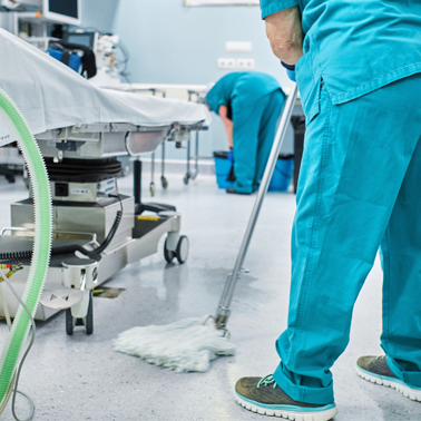 Two hospital staff in teal scrubs, representing premium commercial cleaning services, clean a medical room. One mops the floor as the other tends to equipment near the bed. The setting suggests a sterile environment maintained by expert hands.