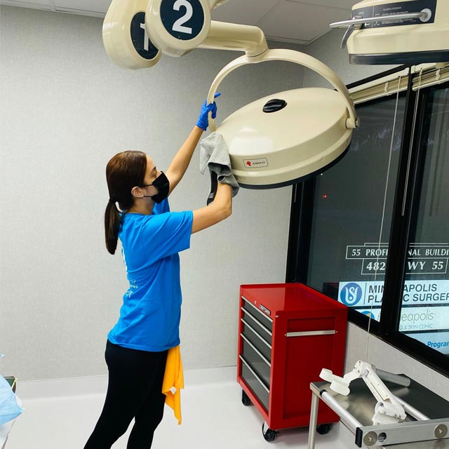 A person in a blue shirt and black mask meticulously cleans a large overhead medical light, showcasing the precision of commercial cleaning services. A red storage cart and windows with signage form the backdrop as they hold a cloth and wear gloves, exemplifying Latin Group Commercial Cleaning's expertise.