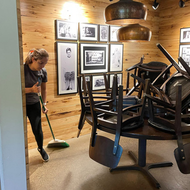 A person from Latin Group Commercial Cleaning is sweeping the floor in a wooden-paneled room with framed photos on the walls. Several stacked chairs are placed on round tables, and ceiling lights illuminate the space.