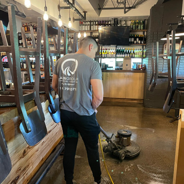 A person in a gray shirt operates a floor cleaning machine, showcasing the efficiency of commercial cleaning services at work. In the restaurant's serene backdrop, chairs are stacked upside down on tables, and shelves stocked with bottles suggest a thorough touch from Latin Group Commercial Cleaning.