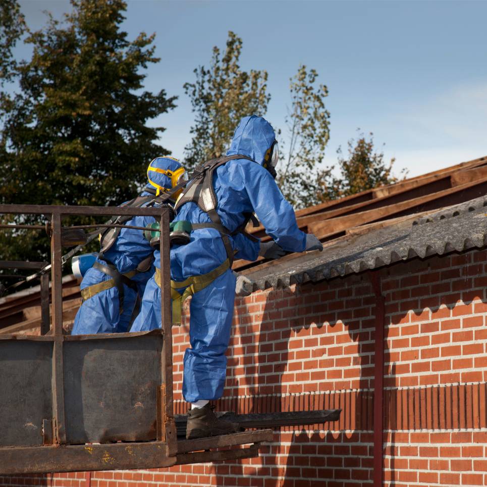 Two workers in blue hazmat suits and helmets are on a lift, expertly conducting commercial cleaning on the sloped roof of a brick building. They are surrounded by trees under a clear sky.