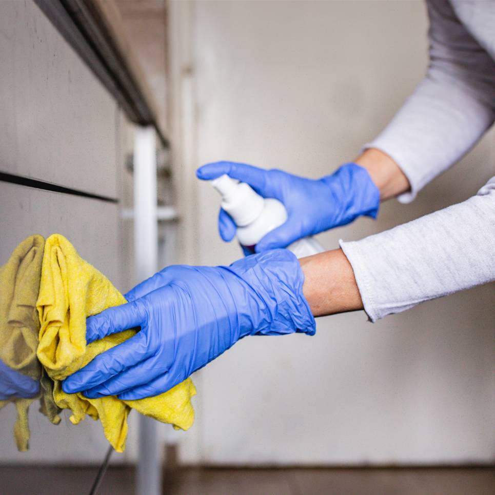 A person wearing blue gloves uses a yellow cloth and a spray bottle to meticulously clean a surface, reflecting the high standards of medical facility cleaning services.