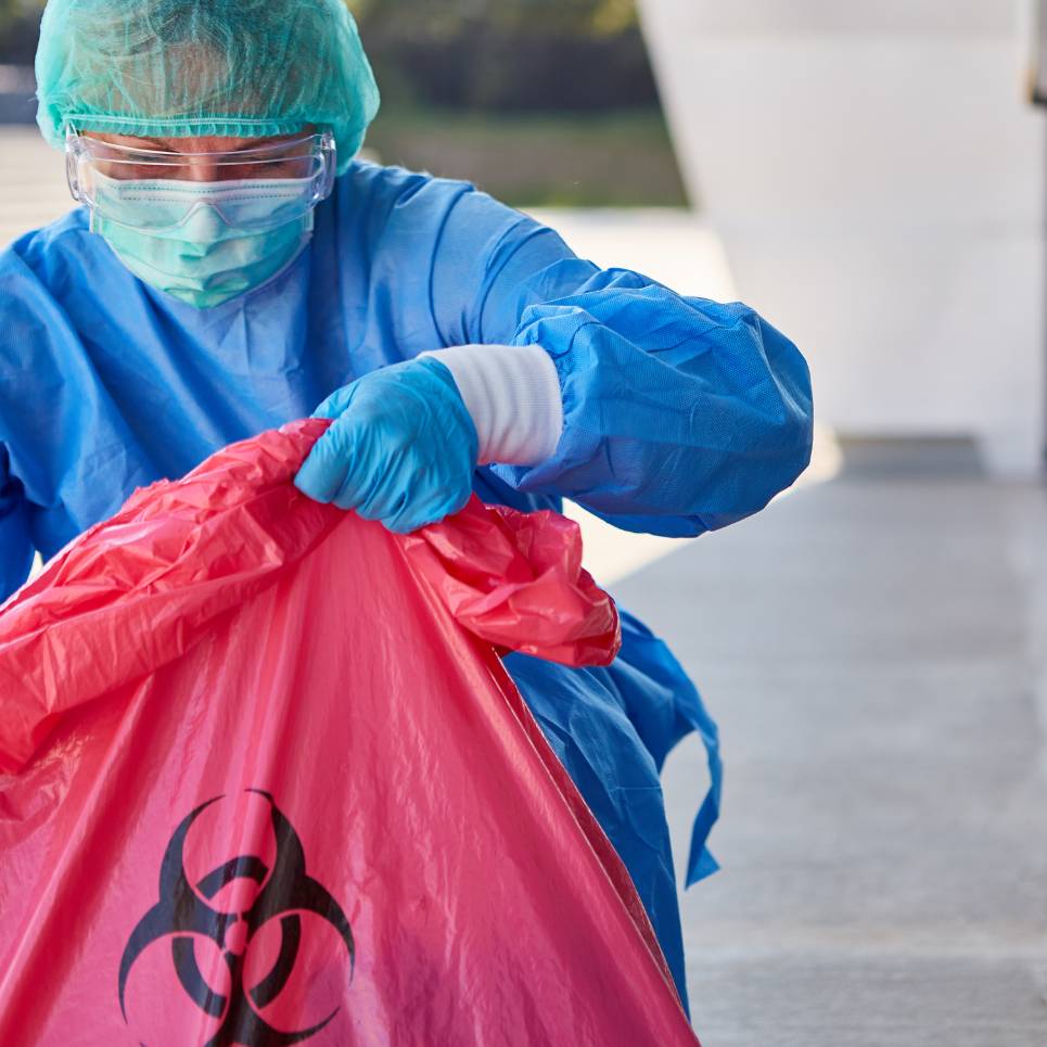 A healthcare worker in protective gear, including a face shield, mask, gloves, and surgical gown, handles a red biohazard bag outdoors as part of medical facility cleaning services.