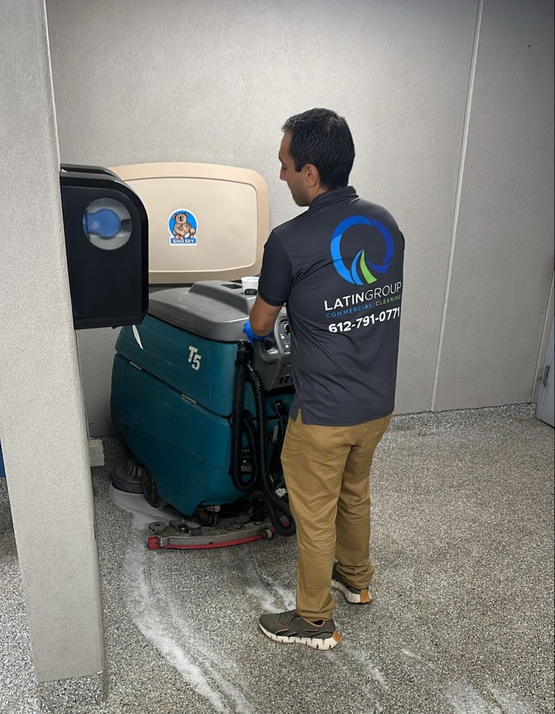 A person operates an industrial cleaning machine in a room. The floor, partially cleaned, glistens with soap suds. Wearing a black shirt adorned with a logo and contact number, they skillfully maneuver the equipment to ensure spotless results.