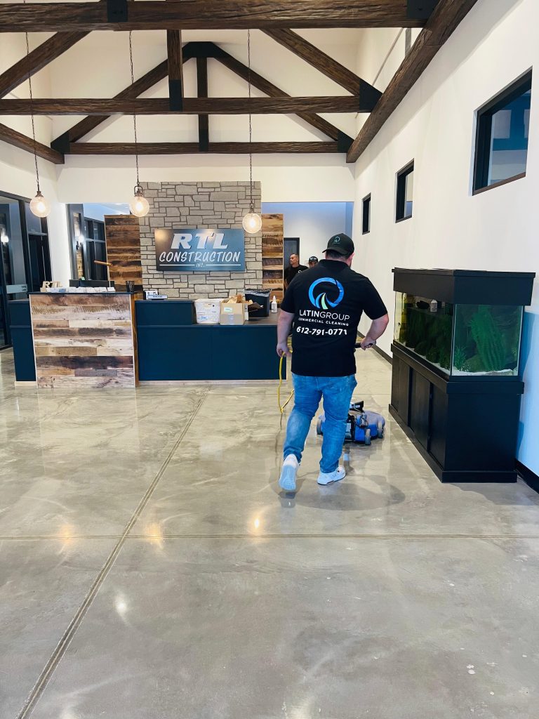 A person wearing a black shirt with a company logo operates an industrial cleaning machine in a modern office lobby. The space features wooden beams, a reception desk with the "RTL Construction" sign, and a large aquarium.