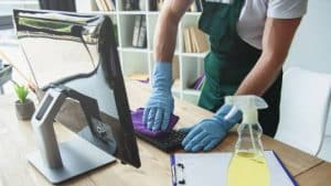 A person wearing blue gloves and a green apron is professionally cleaning a computer keyboard with a purple cloth, ensuring it is thoroughly sanitized. A spray bottle and clipboard are on the desk, beside the computer monitor. Shelves and natural light complete the organized setting.