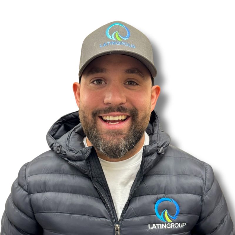 A smiling man wearing a gray cap and a black jacket, both featuring the Latin Grup Commercial Cleaning logo in a blue and green circular design. The background is plain white.