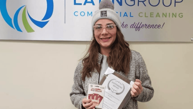 A woman wearing glasses and a beanie smiles while holding an Outback gift card and a certificate. She stands in front of a wall sign that reads "Lawn Group Commercial Cleaning.