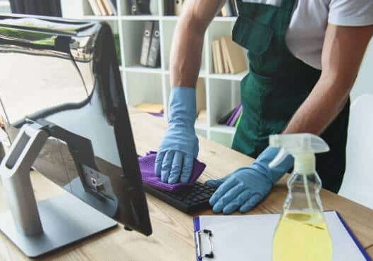 A person wearing blue gloves and a green apron is professionally cleaning a computer keyboard with a purple cloth, ensuring it is thoroughly sanitized. A spray bottle and clipboard are on the desk, beside the computer monitor. Shelves and natural light complete the organized setting.